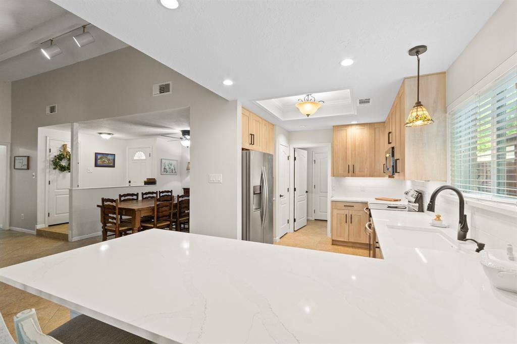 This photo showcases a modern kitchen with a sleek white with grey quartz countertop and stainless steel appliances. It features light wood cabinetry, recessed lighting, and a view into a cozy dining area. The space is bright and inviting, with a window providing natural light.