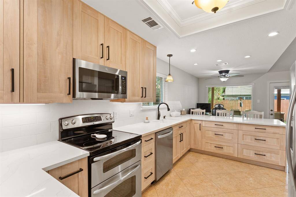 This modern kitchen features light wood cabinetry, stainless steel appliances, and a spacious layout. The room is well-lit with recessed lighting and a central ceiling fan. A large window provides natural light, and the open design includes a breakfast bar with seating, perfect for casual dining.