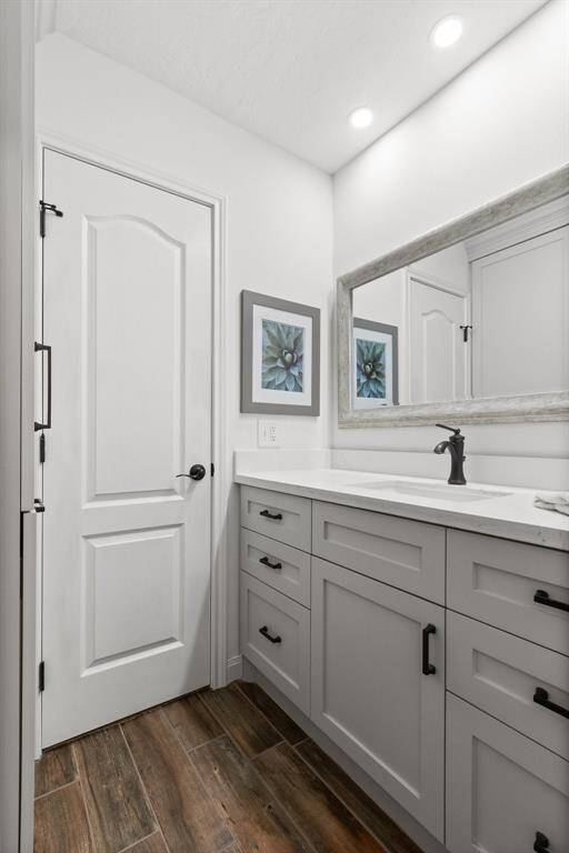 This primary bathroom features a sleek design with white walls, a large framed mirror, and a modern vanity with ample storage. The dark faucet and hardware add a stylish contrast, while wood-look tile flooring adds warmth. Recessed lighting provides a bright, clean ambiance.
