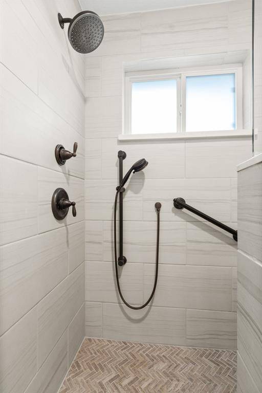 The primary bathroom showcases a modern shower with a sleek design, featuring a rainfall showerhead and handheld fixture. The walls are tiled in a neutral tone, complemented by a herringbone-patterned tile floor. There's a window for natural light and a grab bar for safety.