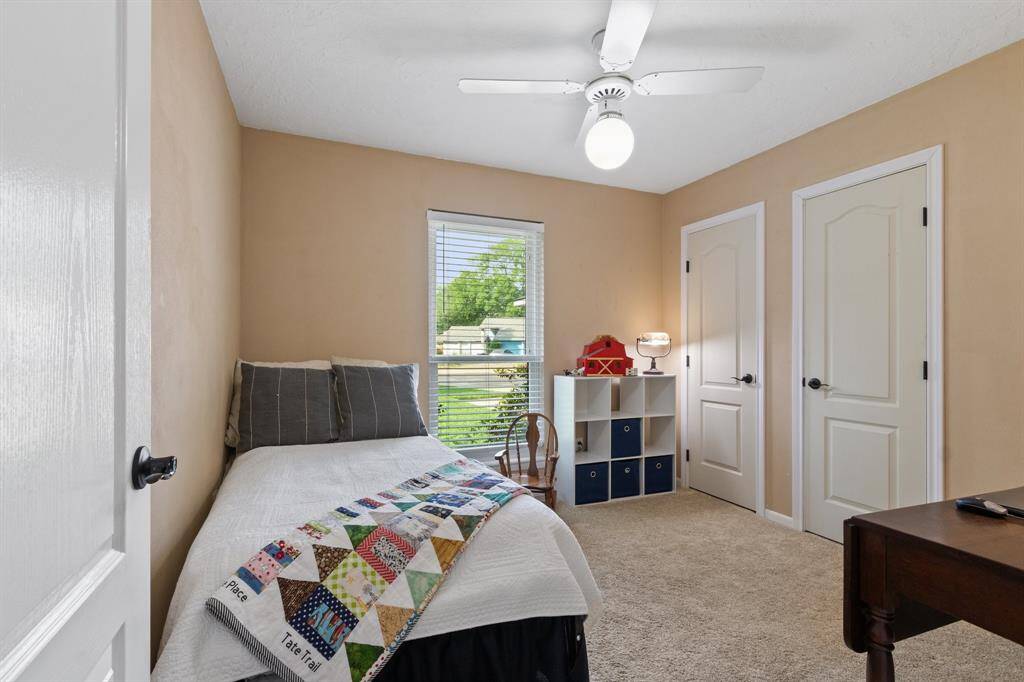 This cozy bedroom features neutral walls, carpeted flooring, and a ceiling fan. It includes a single bed, a small desk, and a window with a view of greenery, offering ample natural light. There are two closet doors for storage, shelving inside, but sellers have the hardware to change it back to hanging clothes closet. Ideal for a child's room or guest space.
