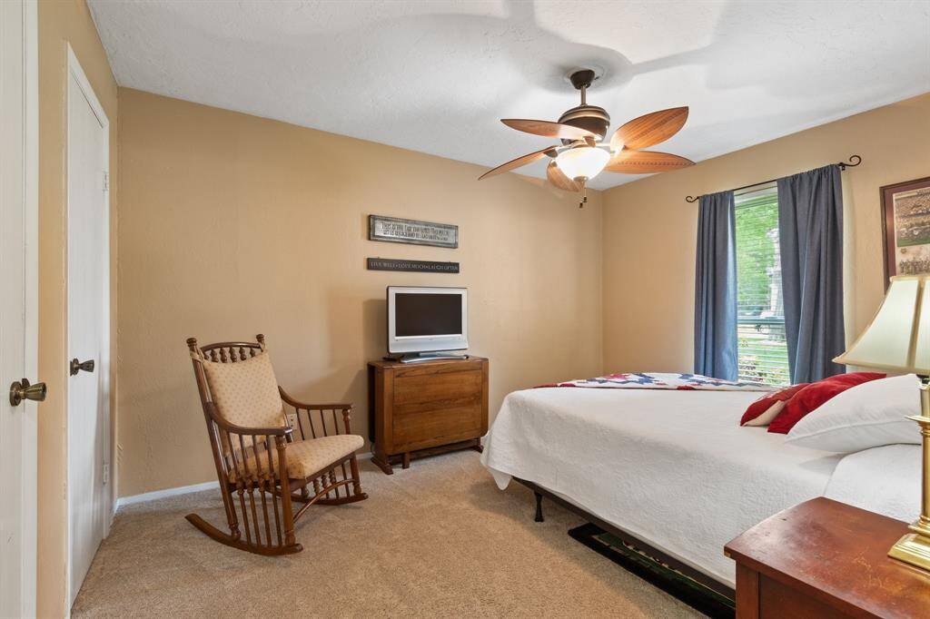 Cozy bedroom with warm beige walls, carpeted floor, and a ceiling fan. Features a window with blinds and curtains, perfect for relaxation.