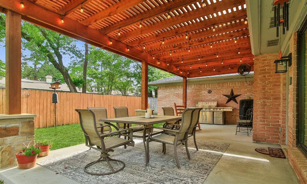 The back patio showcases a charming outdoor space with a wooden pergola. There's plenty of space for a family sized table with chairs, perfect for dining and entertaining. The area can easily accommodate a built-in grill and is surrounded by a privacy fence with lush greenery in the background, providing a cozy and inviting space for relaxation.