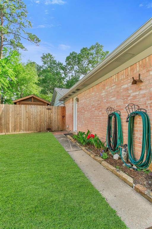 The side yard with a well-maintained lawn and a flower bed along a brick wall. There are garden hoses neatly hung up and a wooden privacy fence with lush trees in the background, creating a serene outdoor space.