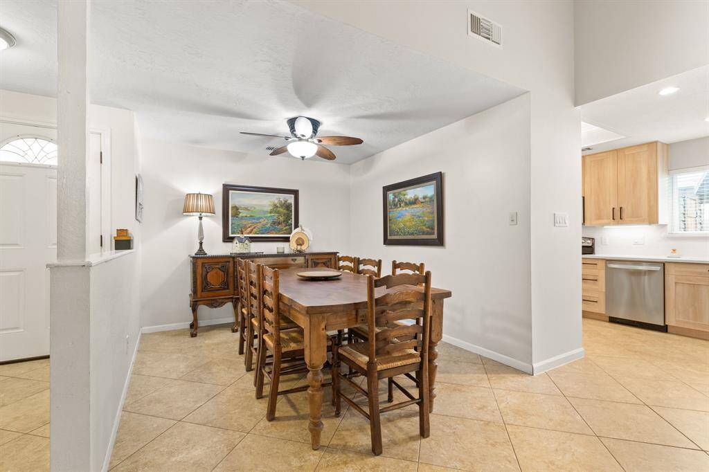 A cozy dining area warmly lit by a ceiling fan. The space has a view into a modern kitchen with light wood cabinets and stainless steel appliances. The room has tiled flooring and an open, inviting layout.