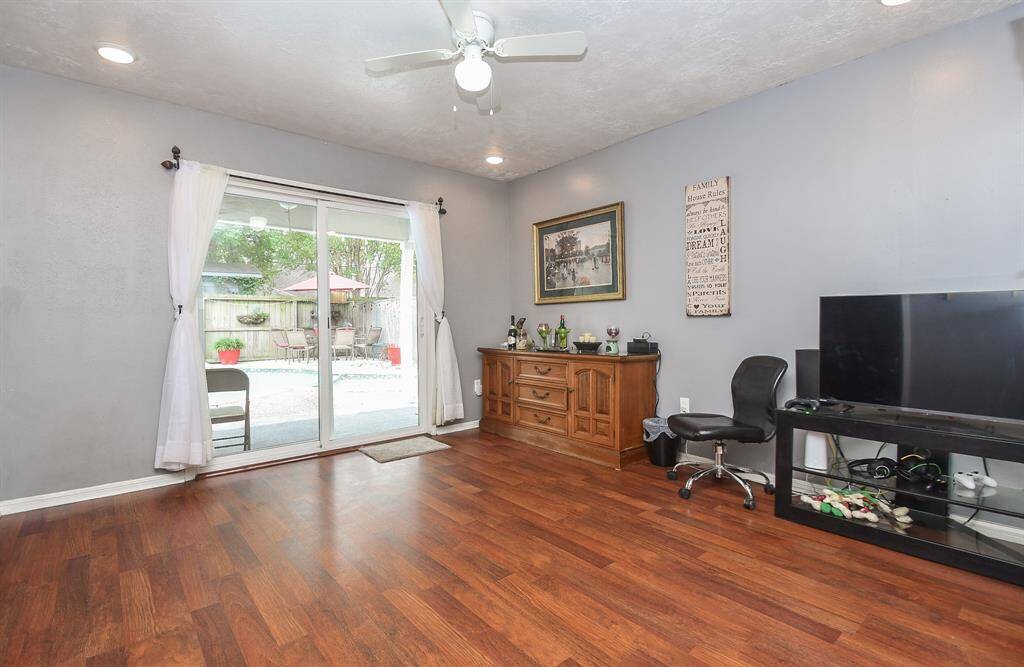 This is a cozy living space featuring laminate wood flooring, neutral wall colors, and ample natural light from a large sliding glass door leading to an outdoor area. The room is equipped with a ceiling fan and has enough space for furniture and entertainment setups.