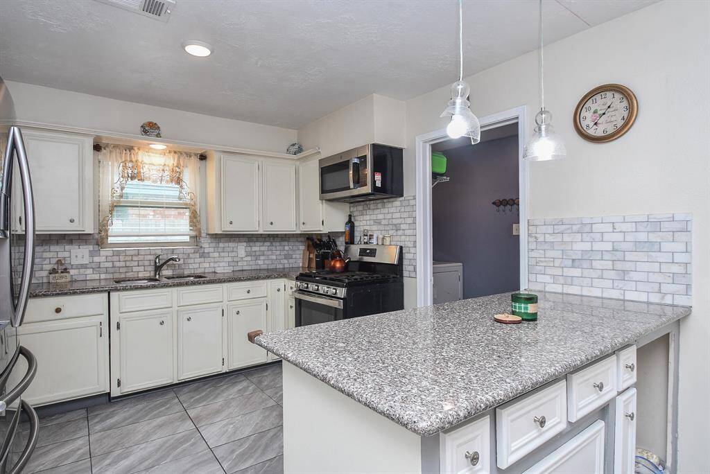 This kitchen features white cabinetry, granite countertops, a subway tile backsplash, modern pendant lights, and stainless steel appliances, with a neutral color scheme and tiled flooring.