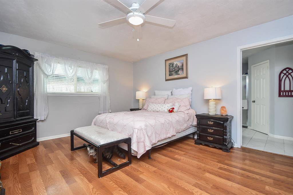 This is a cozy bedroom featuring hardwood floors, a ceiling fan, natural light from a window, and a neutral color scheme.