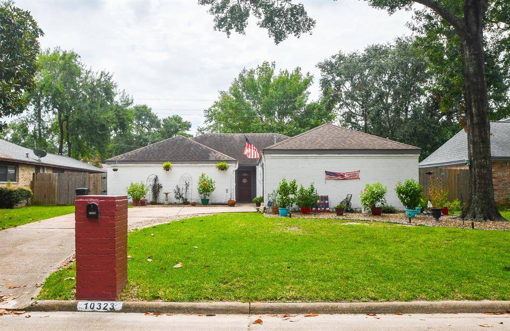 This is a single-story, home with a well-maintained lawn and a brick mailbox. The house features a two-car garage, a peaked roof, and is adorned with an American flag. There are mature trees on the property, providing shade and a cozy neighborhood feel.