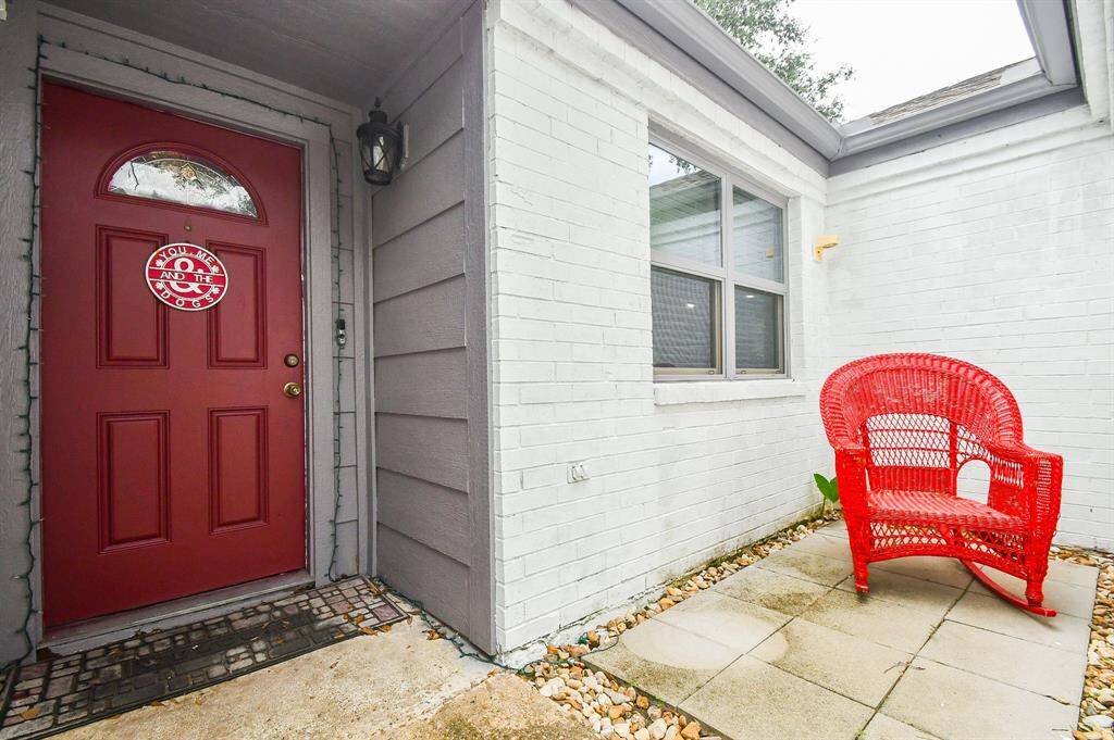 This is a cozy entryway featuring a bold red door with a decorative glass insert and a matching red wicker chair, complementing the home's white painted brick exterior. The space is inviting and offers a small sitting area by the window.