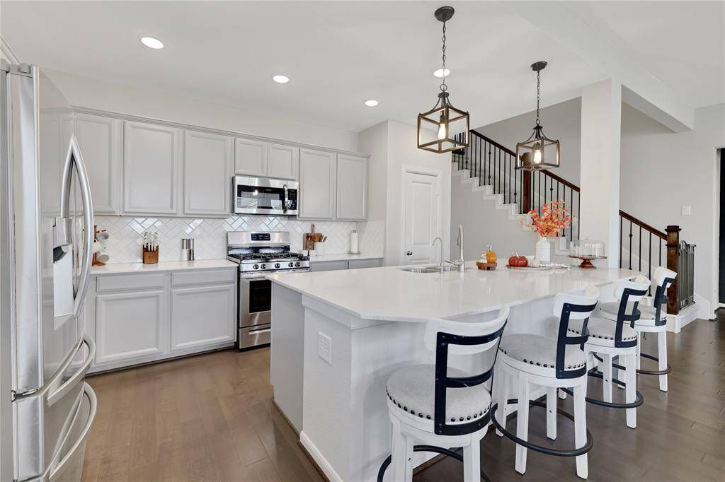The spacious, modern kitchen features white cabinetry, stainless steel appliances, a large island with bar seating, and elegant pendant lighting, with a view of a staircase leading to the upper level.