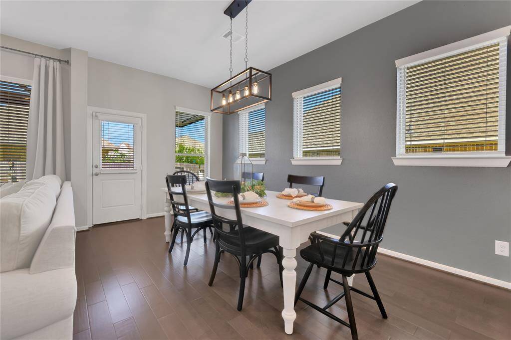 This is a bright, modern dining area, featuring large windows with blinds, sleek lighting, and easy access to the living room area. The space has neutral wall colors and tiled flooring.