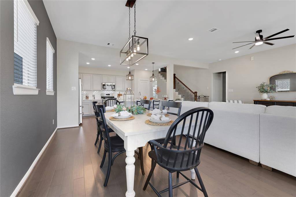 This is a spacious and modern open-concept living space featuring a dining area with a stylish light fixture and a white and gray color palette, The room is well-lit with natural light and recessed lighting,  contributing to an airy and inviting atmosphere.
