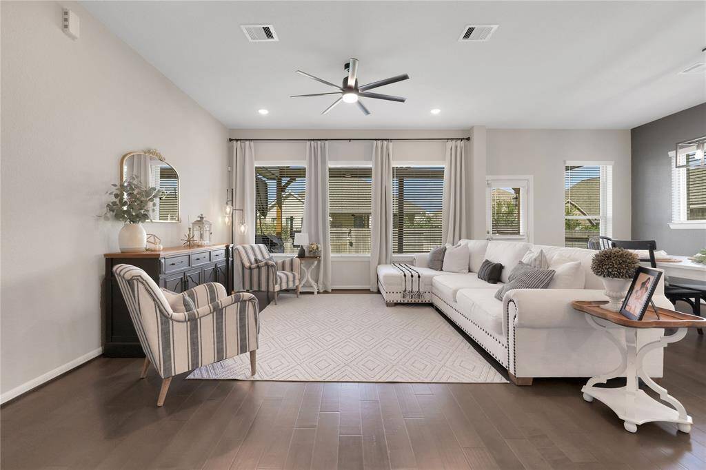 The living room features large windows with blinds, a neutral color scheme, and hardwood floors. with ample space for seating and entertaining.