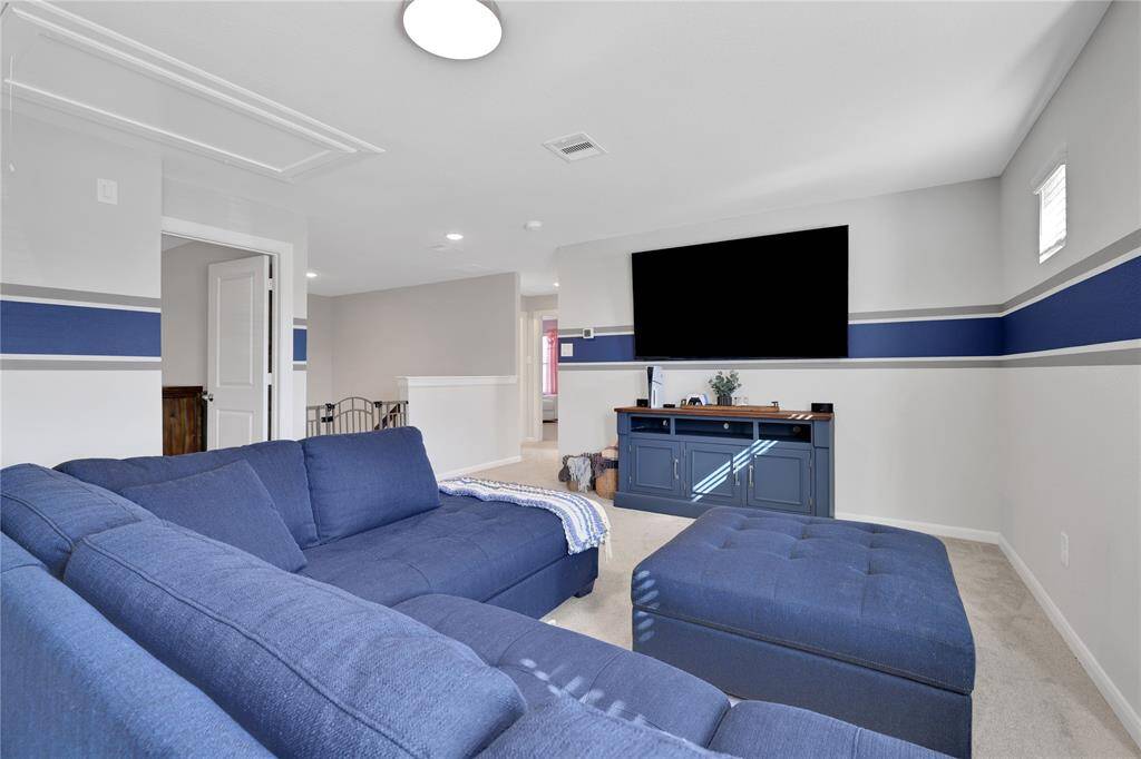 This is a spacious, modern game room with a blue and white color scheme. The room is well-lit with natural light and recessed ceiling lights, and it includes an open doorway leading to adjacent bedrooms.