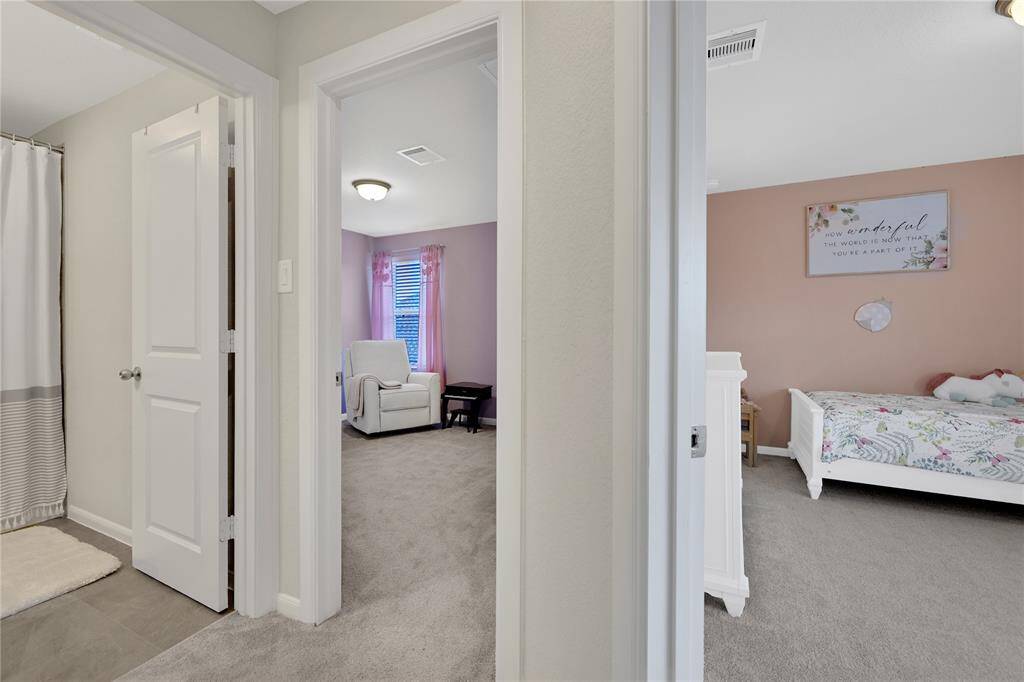 This is a view into a cozy, well-lit bedroom with soft pink walls and a contrasting accent wall, featuring plush carpeting, and a glimpse of an adjoining bathroom.