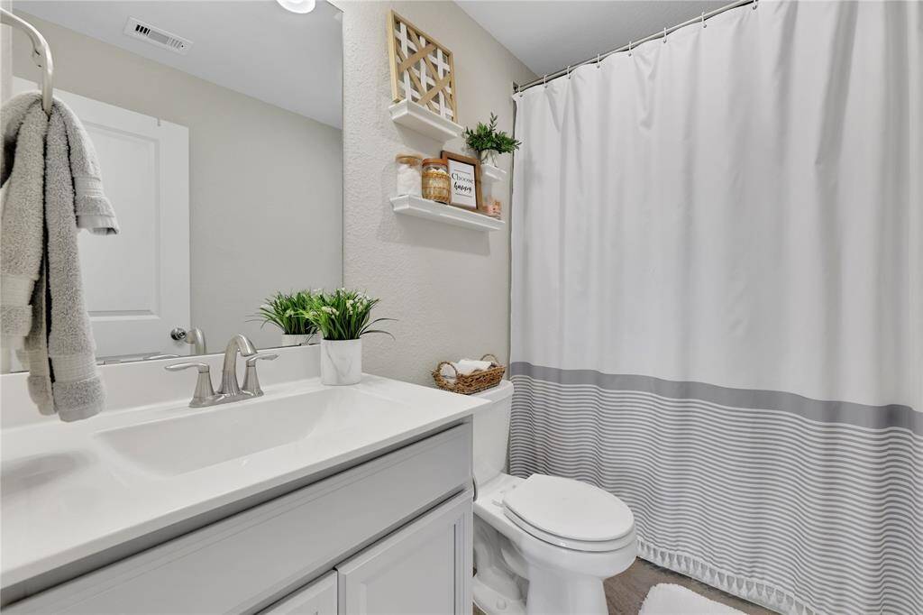 This is a modern and clean bathroom featuring a white vanity with a large mirror, brushed nickel fixtures, and ample storage space. The space is accented with decorative touches like wooden shelving and a gray-striped shower curtain that adds a touch of elegance.