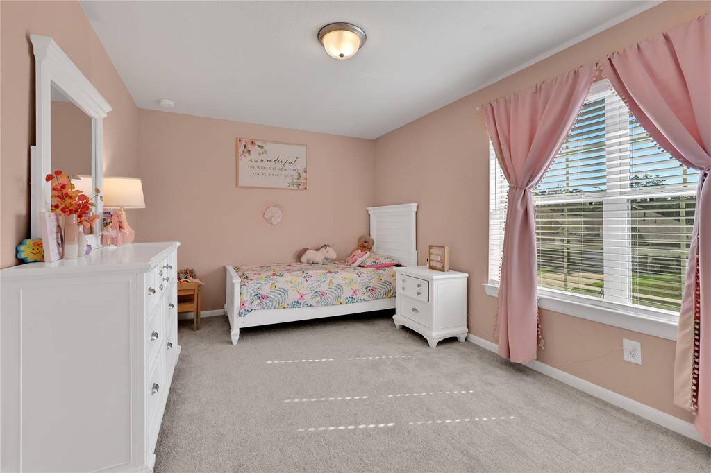 This is a well-lit, cozy bedroom with soft pink walls, a large window, and neutral carpeting.