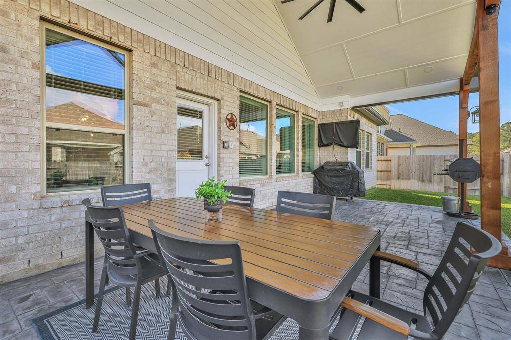 This is a spacious and inviting covered outdoor patio. The area is complemented by a neat stone floor, a ceiling fan for comfort, and large windows that provide a view into the house. The patio opens up to a well-maintained lawn, bordered by a wooden privacy fence.