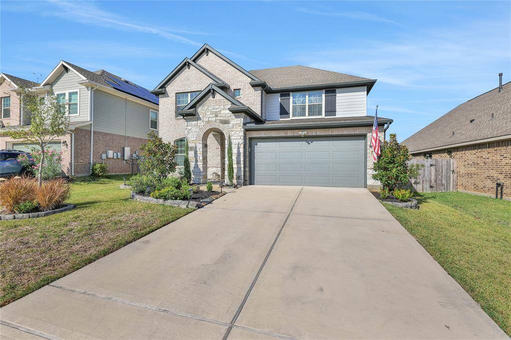 This is a two-story suburban home featuring a stone and siding facade, a two-car garage, and a manicured front yard with a variety of shrubs and a small tree. The house has a welcoming entrance with an arched doorway. The driveway provides ample space for parking, The neighborhood is well-maintained .