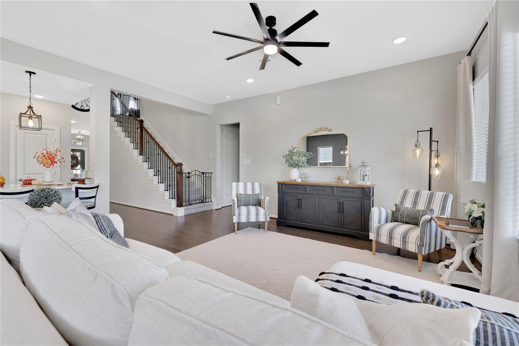 The well-lit living room with a neutral color palette, featuring an elegant staircase, and a modern ceiling fan; The room flows seamlessly into the adjacent dining area, providing a cohesive open-plan living space.