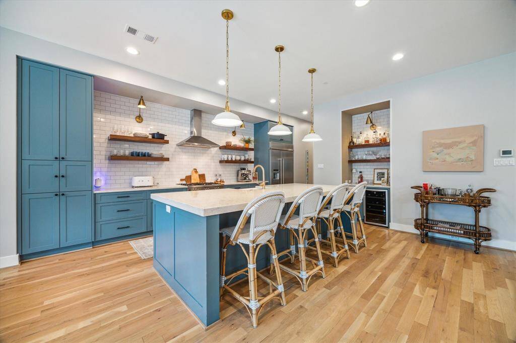 This kitchen is a showstopper, featuring an oversized Silestone island lighted by triple pendants from Visual Comfort. Custom inset Shaker-style cabinetry by J-Kraft Inc., a polished ceramic subway tile wall, and floating wooden shelves highlighted by brass sconce lighting add to its stunning appeal.