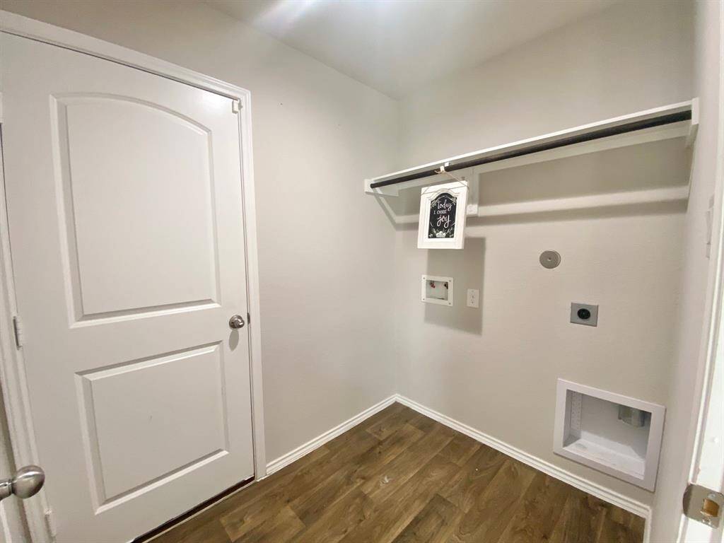Laundry room with wood flooring, a single door, and hookups for a washer and dryer. There's a shelf for storage and neutral-colored walls, creating a clean and functional space.