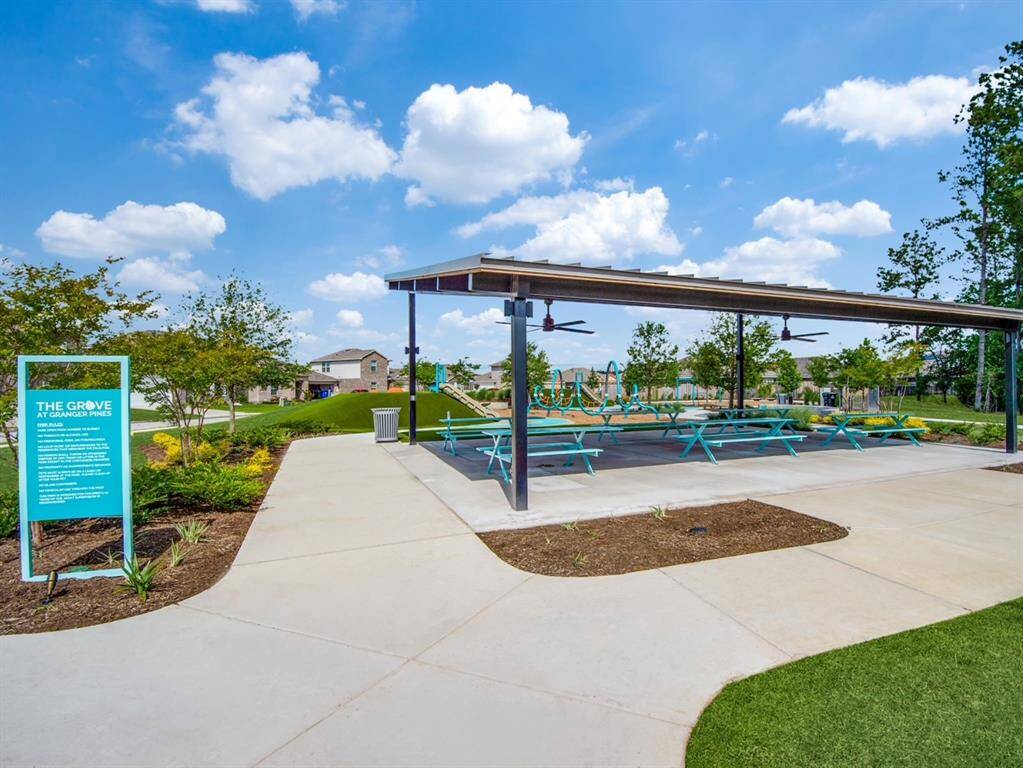 Community park area featuring a covered pavilion with picnic tables, ceiling fans, and a nearby playground. The setting is surrounded by landscaped greenery, with a clear path leading through the space. Ideal for outdoor gatherings and picnics and birthdays