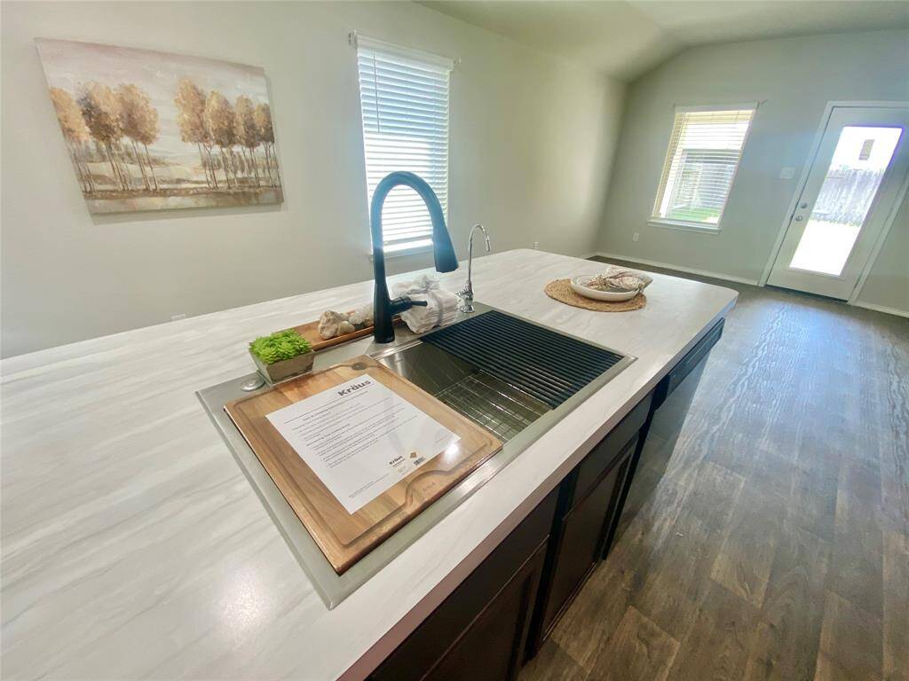 Modern kitchen with a large island featuring a sleek sink and faucet. Bright, natural light fills the space through large windows and a glass door, complementing the wood flooring and neutral walls.