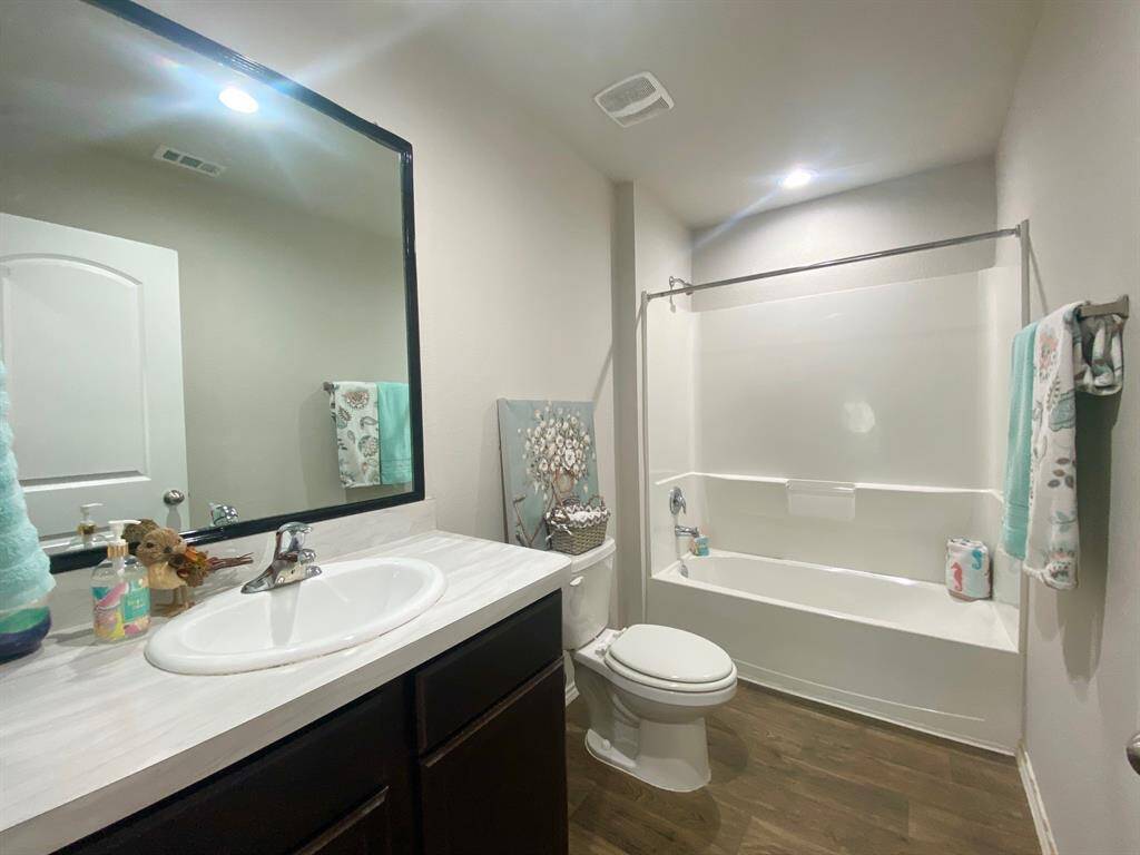 This is a bright, modern bathroom featuring a large mirror, a vanity with ample counter space, a bathtub with shower, and wood-style flooring.