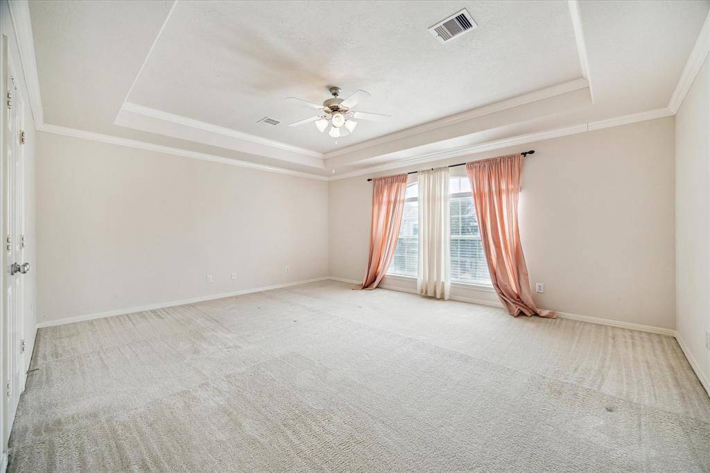 The Primary Bedroom offers a spacious layout with a soothing neutral interior, elegant tray ceilings with crown molding, and picturesque views of the front yard, creating a peaceful and refined retreat.