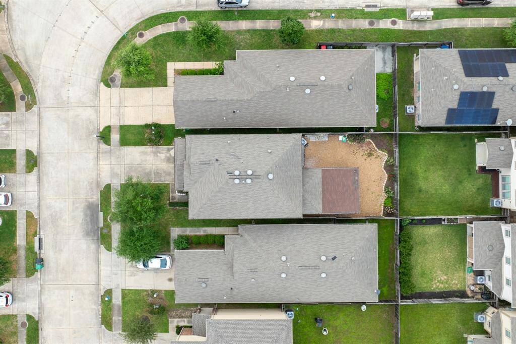 This aerial photo showcases a suburban neighborhood featuring well-maintained lawns and driveways, and is surrounded by greenery. Streets are wide and lined with trees, offering a peaceful, residential atmosphere.