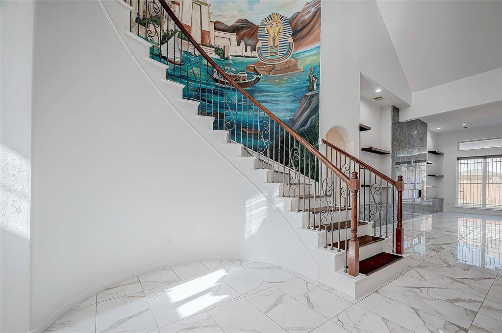 Upon entry, a curved staircase with wood steps, wrought iron spindles, and a stunning chandelier creates a grand impression, accented by elegant wall painting.