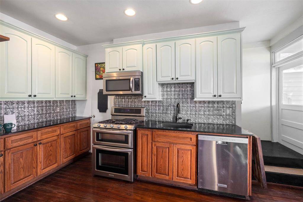 This kitchen features a charming blend of modern and rustic styles with stainless steel appliances, black countertops, and a decorative tin backsplash. The cabinetry combines white and wood finishes, and the space is brightened by recessed lighting.