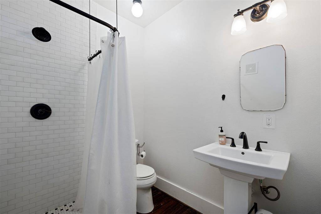 This bathroom off the downstairs bedroom features a sleek design with white tile walls, a shower with a curtain, a pedestal sink, and a modern black faucet. The space is well-lit with mounted light fixtures, creating a clean and bright atmosphere.