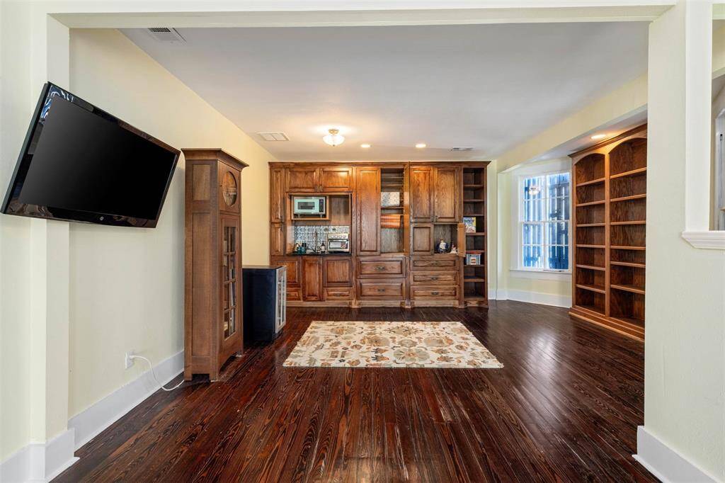 This room features rich hardwood floors, kitchenette, wrap station and built-in wooden cabinetry with ample storage space. The area is well-lit with recessed lighting and a large window, and a mounted TV adds modern convenience.