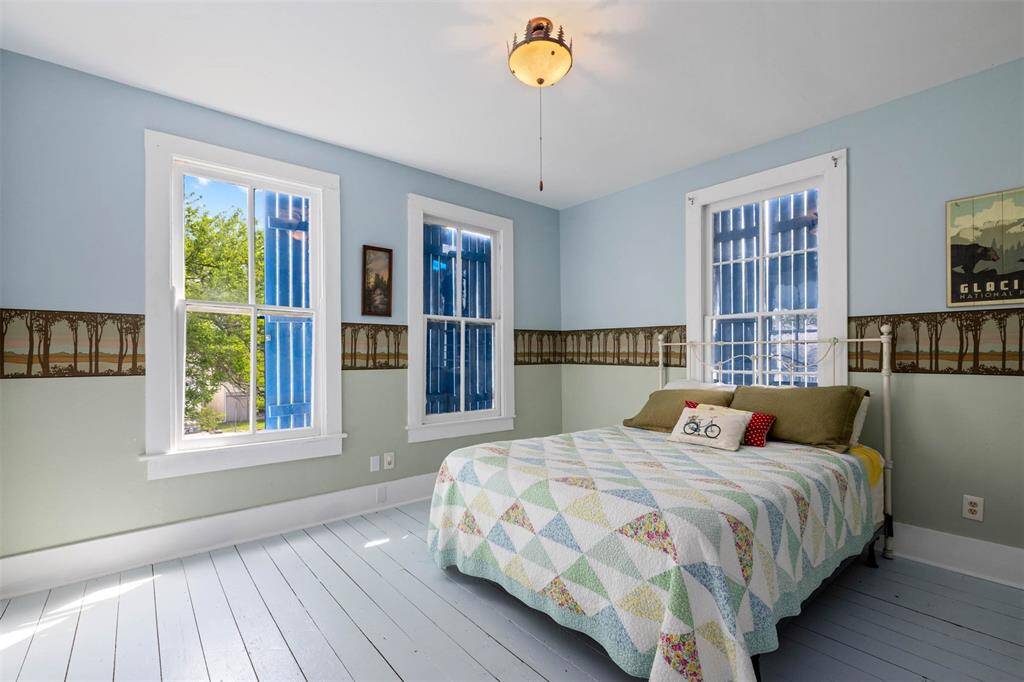 Bright and cozy bedroom featuring light blue walls, a ceiling fan, and three large windows providing abundant natural light and complemented by a scenic tree border on the walls and fun light fixture on the ceiling.