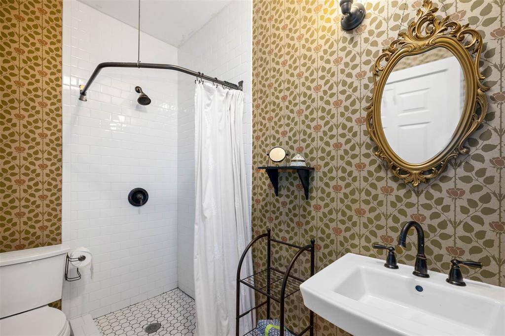 This bathroom features a walk-in shower with white subway tiles, a classic white sink, and a toilet. The walls are adorned with a floral-patterned wallpaper, and there's an ornate gold-framed mirror above the sink, adding a touch of elegance.