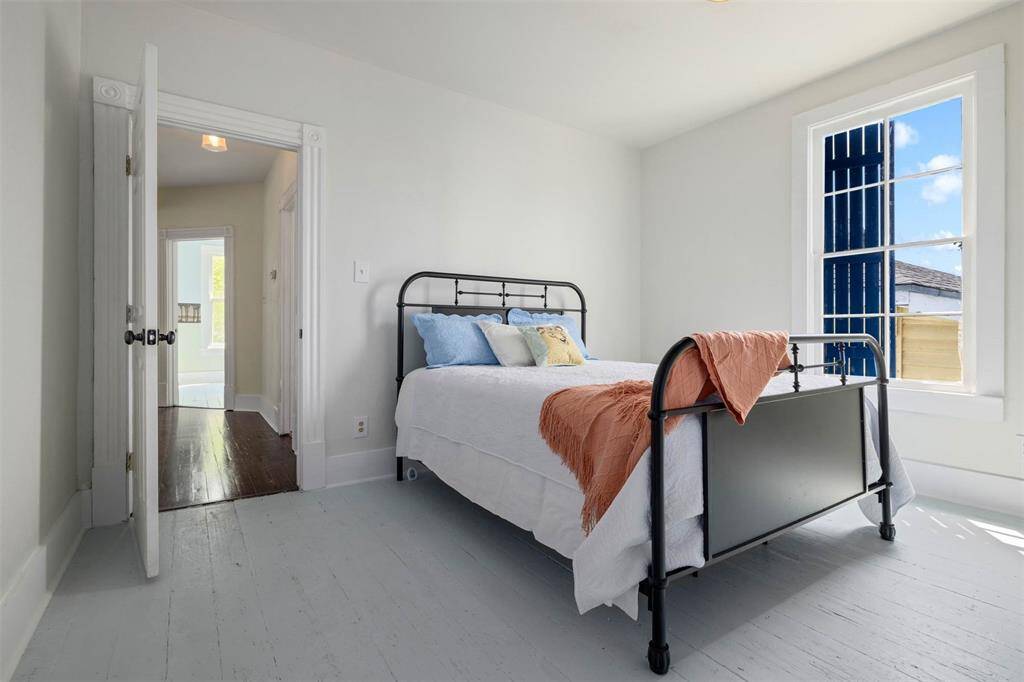 This bright bedroom features a vintage metal bed frame, light-colored walls, hardwood floors, and a window allowing natural light to fill the space. The open doorway connects to a hallway with additional natural light.
