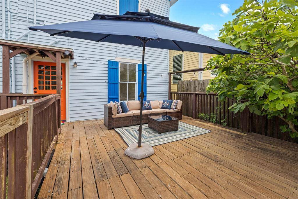 This photo features a charming outdoor deck with wooden flooring, a cozy seating area under a large blue umbrella, and a vibrant orange door. It’s an inviting space perfect for relaxation or entertaining, surrounded by greenery and offering a touch of color with blue window shutters.