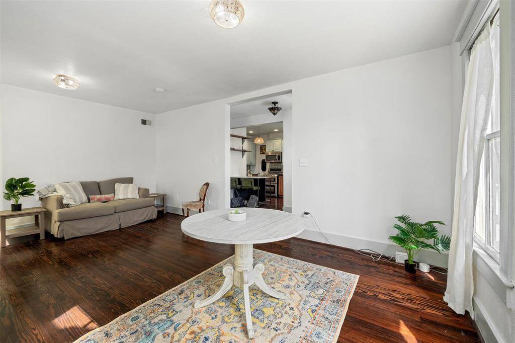 Bright living area with hardwood floors, neutral walls, and ample natural light. Includes a cozy seating area, dining area and glimpses of a modern kitchen through an open doorway.