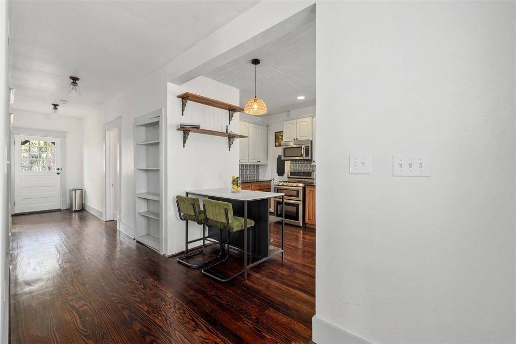 This bright and inviting home features a sleek kitchen with modern appliances and an island with seating. The open hallway leads to a well-lit entryway. Hardwood floors and built-in shelving add charm and functionality.
