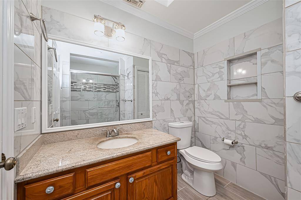 Upstairs bathroom features elegant marble-patterned tile walls, a granite countertop with a wooden vanity, and a large mirror. It includes a new toilet and a shower with stylish tile accents, providing a sleek and functional space.