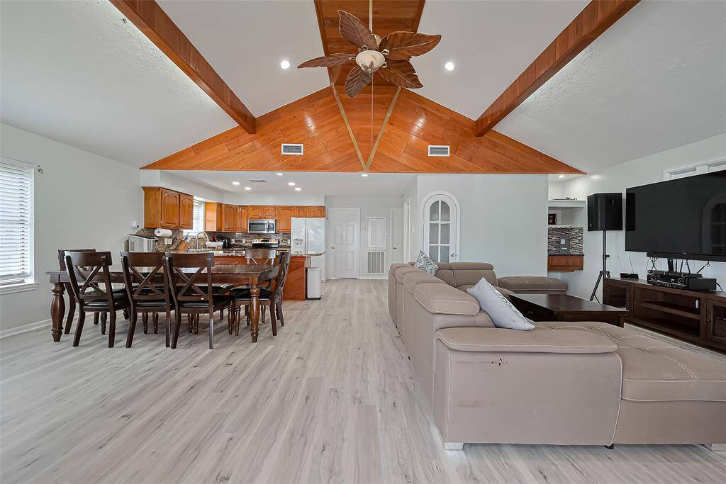 Spacious open-concept living area features a vaulted ceiling with wood accents and a dining area that flows into a modern kitchen with wooden cabinets and stainless steel appliances. New laminate flooring and neutral tones create a warm, inviting atmosphere.