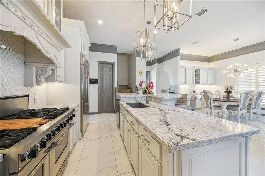 This elegant kitchen features a spacious island with a sink and high-end stainless steel appliances. The open layout flows into a dining area with stylish lighting and ample natural light, creating a bright and inviting space.