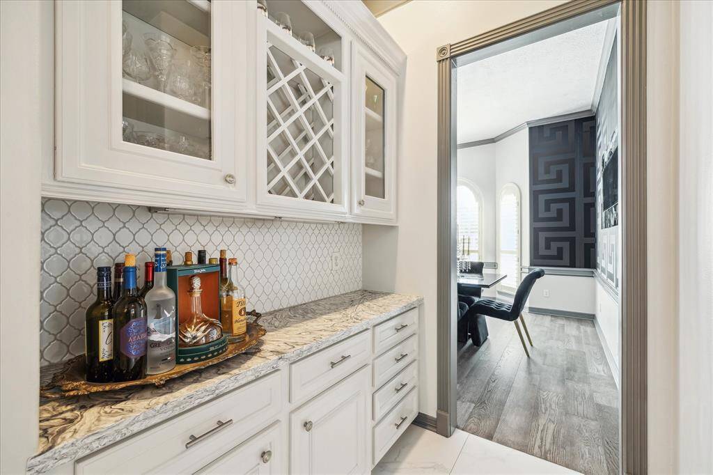 This photo showcases a stylish butler's pantry with white cabinetry, countertops, and a wine rack. It leads into a bright dining area with elegant decor and wood flooring.