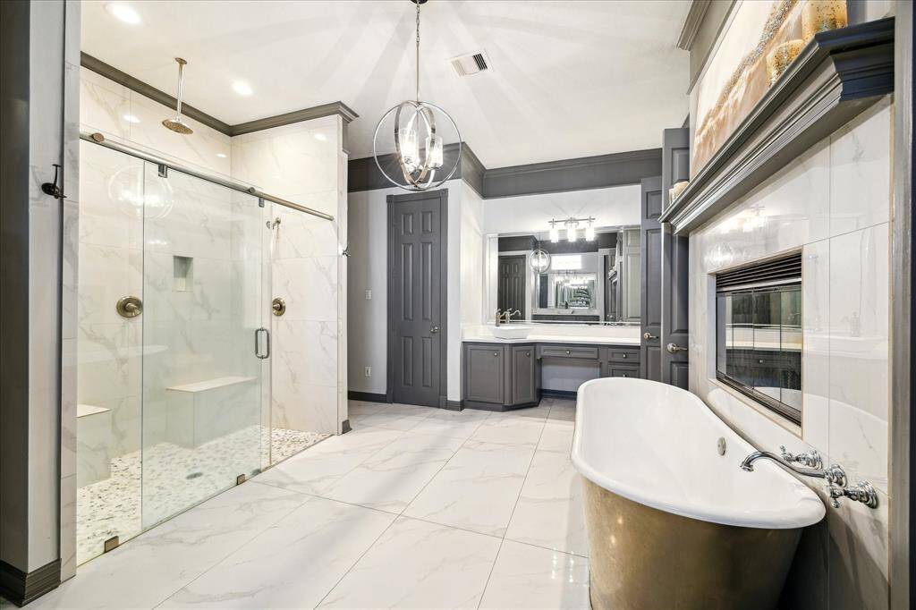 This luxurious bathroom features a large walk-in shower with glass doors, a freestanding tub, and a dual-sink vanity. It has elegant marble flooring, a modern chandelier, and a cozy fireplace, creating a spa-like atmosphere; also featuring dual water closets.