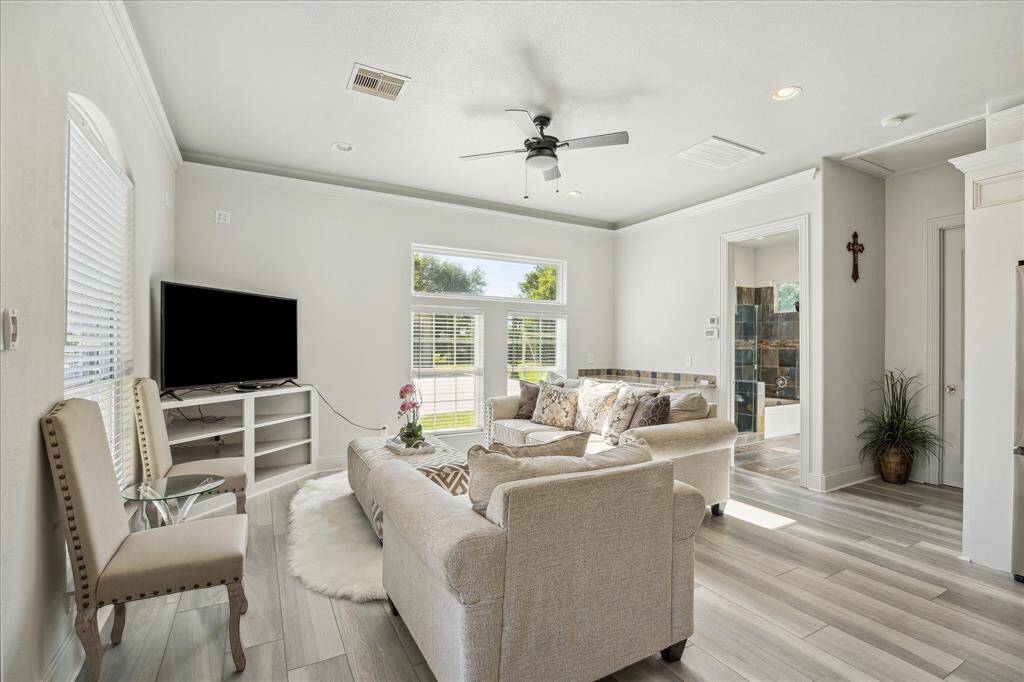 This bright living room features a stylish arched door with ornate ironwork, modern ceiling fan, and large windows for natural light. It includes comfortable seating, light wood flooring, and a wall-mounted TV, creating a cozy and inviting space.