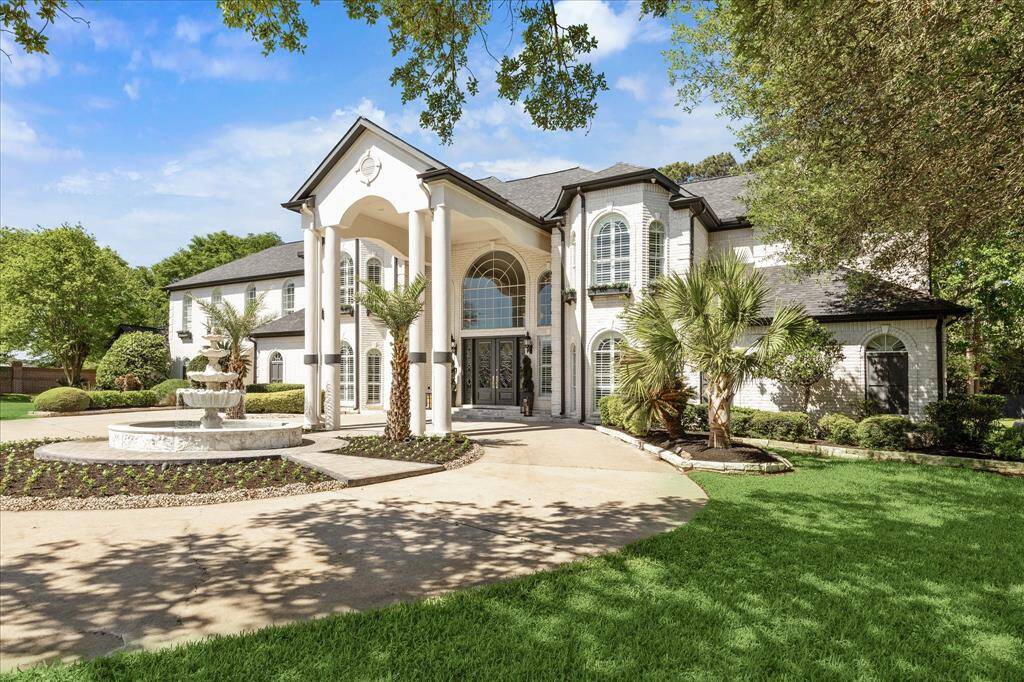 This photo showcases a grand two-story home with a striking entrance featuring tall white columns and large, arched windows. The landscaped front yard includes a circular driveway, a fountain, and lush greenery.