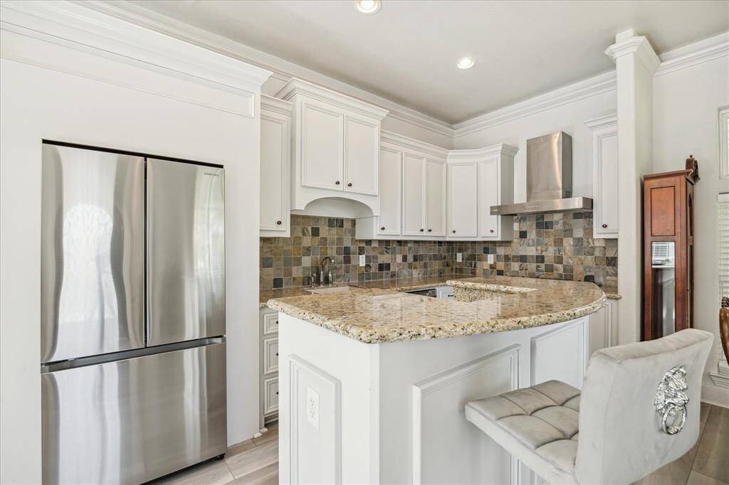 This bright kitchen features white cabinetry, a stylish tiled backsplash, and stainless steel appliances. It includes a cozy dining area with an elegant chandelier and a glass-top table, complemented by large windows that let in plenty of natural light.